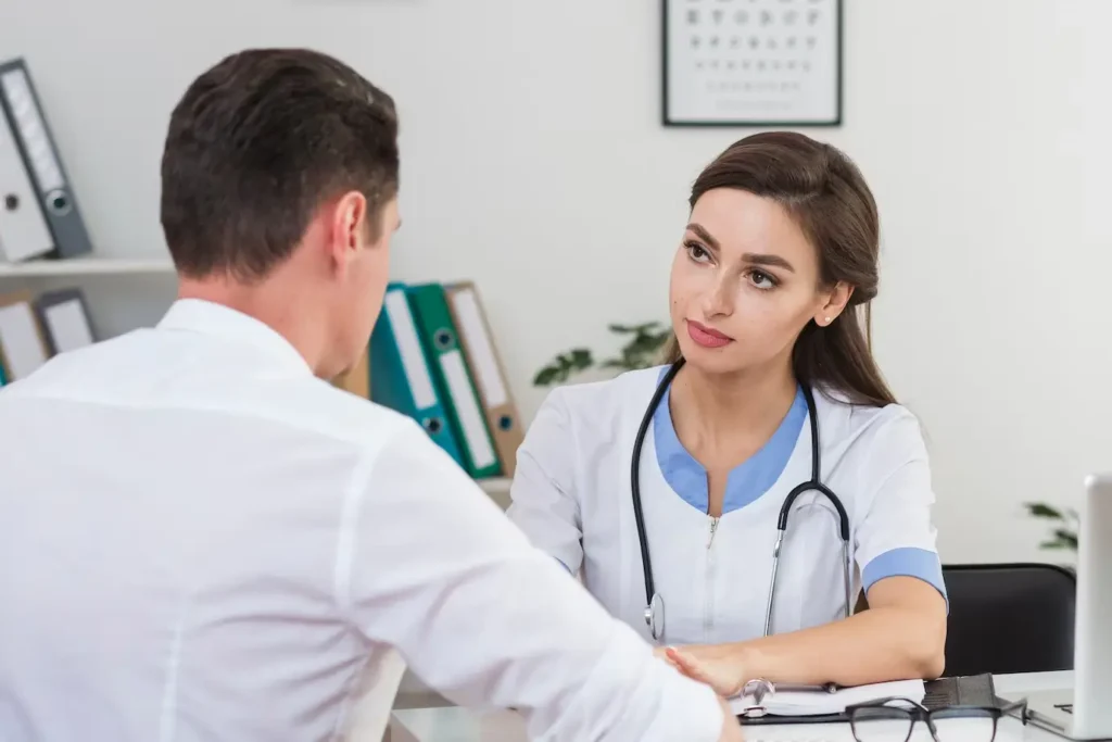A female doctor in a white coat with a stethoscope is sitting at a desk and listening to a male patient who has his back to the camera.