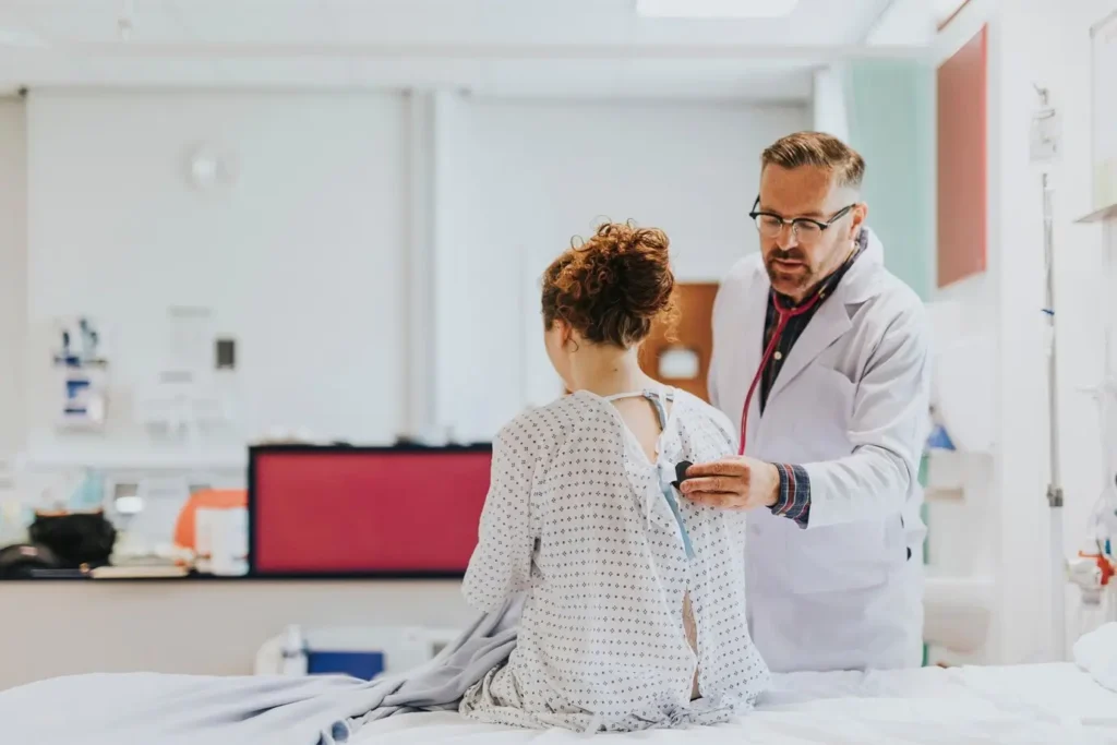 A male doctor is using a stethoscope to listen to the back of a female patient who is sitting on a hospital bed.