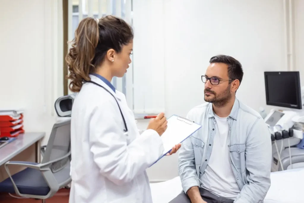 A young female doctor in a white coat with a stethoscope is holding a clipboard and talking to a male patient who is sitting on an examination table.