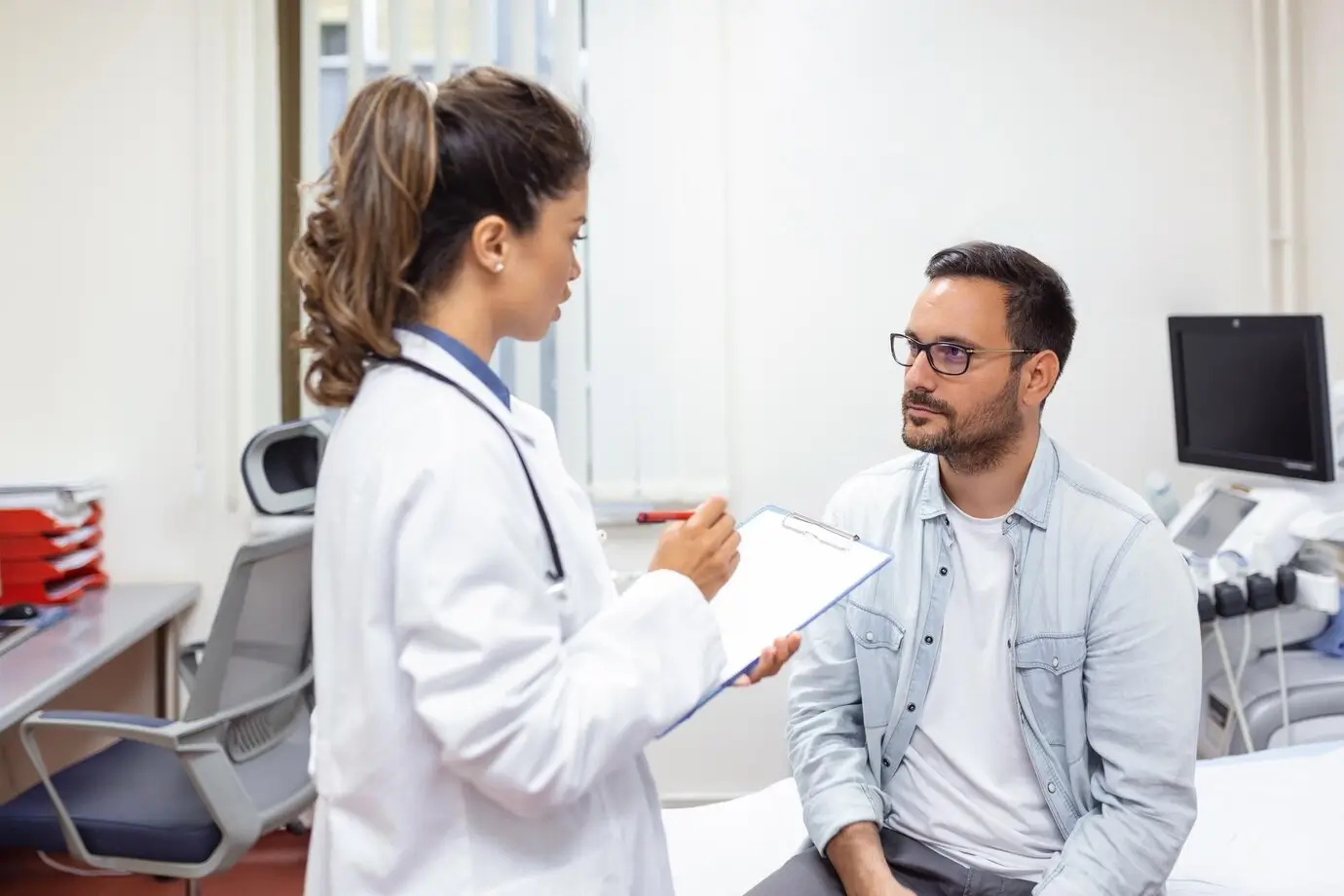 A young female doctor in a white coat with a stethoscope is holding a clipboard and talking to a male patient who is sitting on an examination table.